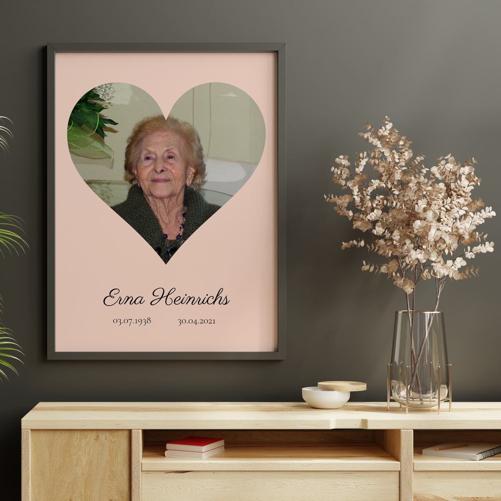 Trauerfoto mit Herzporträt auf Rosa im dunklen Rahmen an grauer Wand über einem Holzsideboard neben Trockenblumen