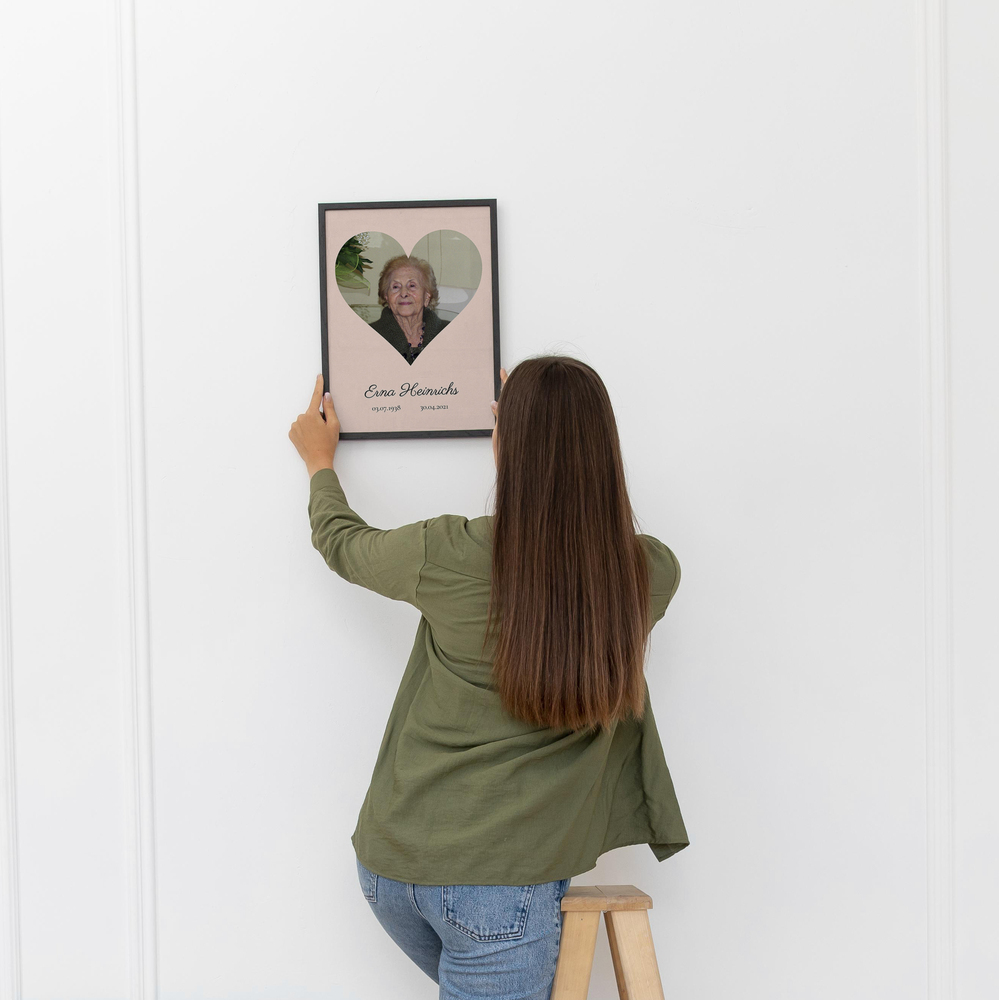 Trauerfoto mit Porträt in Herzform, Namen und Lebensdaten im dunklen Rahmen an einer weißen Wand